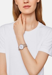 Hellbeige Uhr mit einem runden weißen Ziffernblatt und minimalistischer Gestaltung, ausgestattet mit einem glatten Lederarmband. Das Modell trägt ein weißes T-Shirt.