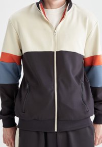 Veste zippée colorée à blocs, avec des panneaux crème, brun foncé, bleu et orange ; comprend des poches zippées sur les côtés et des poignets côtelés.