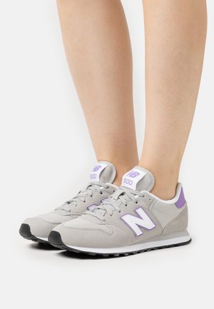 Pieds portant des baskets New Balance 500 grises et violettes avec des semelles intermédiaires blanches et des semelles extérieures noires, sur un fond blanc uni.
