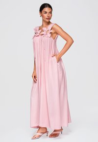 Femme aux cheveux foncés portant une longue robe ample rose pâle avec des détails floraux sur les bretelles et tenant une poche, debout devant un fond uni.