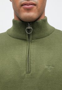 Ζακέτα με φερμουάρ σε ελιά πράσινο χρώμα με ριμπ γιακά και υφή. Διαθέτει ασημένιο φερμουάρ και μικρό λογότυπο στο αριστερό στήθος.