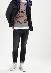 Uomo che indossa un giubbotto imbottito nero, un maglione grigio con scritta rossa, jeans neri e sneakers alte bianche, in piedi contro uno sfondo bianco.