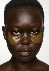 Pelle marrone con capelli corti, indossando patch per gli occhi in gel verde, texture della pelle liscia, espressione neutra, sfondo minimale.