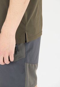 Oliivivihreä t-paita, jossa on lyhyt sivuhalkio, yhdistettynä harmaisiin shortsit, joissa on vetoketjullinen tasku ja kontrastisia kankaita.