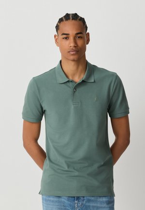 Jeune homme aux cheveux tressés portant un polo vert manches courtes et un jean bleu, debout devant un fond clair uni.