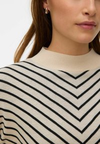 Maglione beige con strisce a chevron nere, caratterizzato da un collo alto a coste. Il modello indossa un piccolo orecchino d'argento curvo. Texture liscia.
