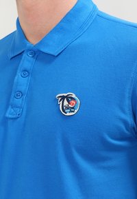 Blaues Poloshirt aus strukturiertem Stoff mit einer Knopfleiste mit vier Knöpfen. Gesticktes Patch mit Palme und "73"-Detail auf der linken Brust.