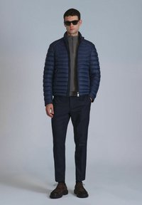 Navy padded jacket with high collar, zip front, and side pockets, worn over a gray top; paired with dark trousers and brown shoes.