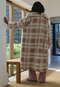 Płaszcz z beżowo-brązowym w kratkę, z dużym kołnierzem, długimi rękawami i miękką teksturą; noszony na jasnoróżowych spodniach, stojąc obok drewniej ławki.