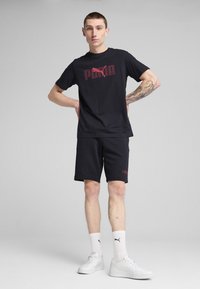 Melnas kokvilnas T-krekls ar sarkano Puma logo, kombinēts ar melniem šortiem. Modelis valkā baltas kedas un potītes zeķes ar Puma logo.