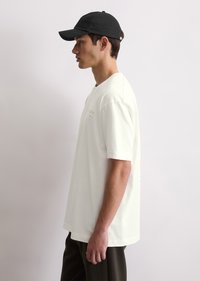 White cotton t-shirt with short sleeves and crew neck, featuring subtle text details on the front and back, paired with a black baseball cap.