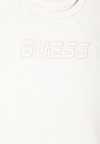 Biała bluza z wypukłym logo "GUESS" w tym samym kolorze, z okrągłym dekoltem i żeberkowanymi detalami na ramionach. Miękka faktura materiału.