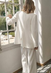 Biały, oversize'owy żakiet o eleganckim fasonie, w połączeniu z szerokimi spodniami. Gładka tkanina i dopasowany krój. Naturalne światło rzuca delikatne cienie.