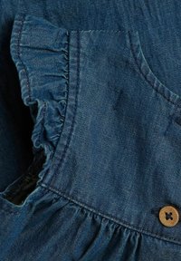 Denim kledingstuk met ruches aan de mouwen, donkerblauwe kleur en een houten knoopaccent op het lichaam.