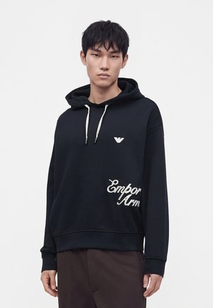 Nuori mies, jolla on musta huppari valkoisilla naruilla ja Emporio Armani -logo, seisoo yksinkertaisen valkoisen taustan edessä.