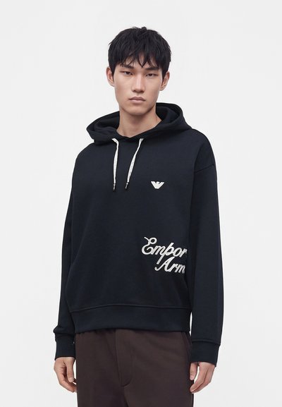 Jeune homme portant un sweat à capuche noir avec des cordons blancs et le logo Emporio Armani, debout devant un fond blanc uni.