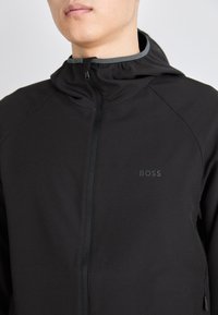 Črna zip-up jakna z visokim ovratnikom, narejena iz gladkega materiala. Ima subtilno znamčenje na prsih in funkcionalen zadrgo.