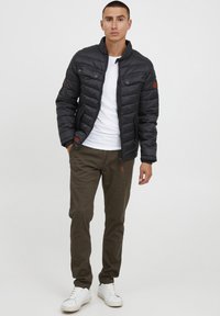 Veste en duvet noire avec un col montant, présentant un design matelassé et des accents bruns, associée à un pantalon vert olive et des baskets blanches.