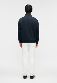 Μπλε σκούρο φούτερ με ψηλό γιακά και ριμπ τελειώματα, σε συνδυασμό με λευκό παντελόνι. Το ύφασμα φαίνεται λείο και ελαφρώς υφασμένο.