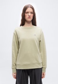 Νεαρή γυναίκα με μακριά ίσια μαλλιά, ντυμένη με ανοιχτό μπεζ φούτερ και σκούρο παντελόνι με φαρδιά μπατζάκια, στέκεται μπροστά σε μια απλή λευκή φόντο.