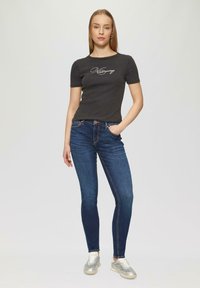 T-shirt noir à manches courtes côtelées avec un design script, associé à un jean skinny bleu foncé et des baskets métalliques argentées sur un fond clair.