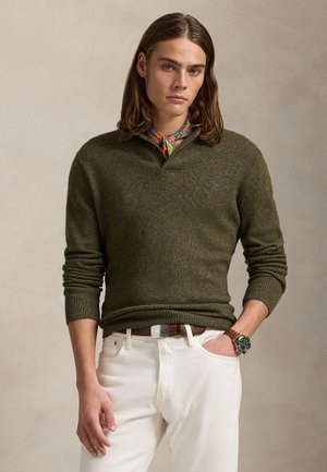 Jeune homme aux cheveux mi-longs portant un pull vert olive, un pantalon blanc, un foulard coloré autour du cou et une montre-bracelet, debout devant un fond neutre.