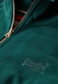 Chaqueta de felpa de color verde azulado con cremallera plateada. Presenta el logotipo bordado de "SuperDry". Textura suave, diseño casual, vista en primer plano.