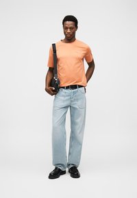 Πορτοκαλί βαμβακερό T-shirt, ανοιχτό μπλε τζιν παντελόνι και μαύρα παπούτσια. Το μοντέλο κρατάει μια μικρή μαύρη τσάντα χιαστί, στέκεται σε ουδέτερο φόντο.