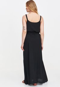 Osoba z długimi blond włosami nosi czarną, bezrękawnikową sukienkę maxi, odsłaniając tatuaż na górnej części ramienia, stoi na jednolitym tle.