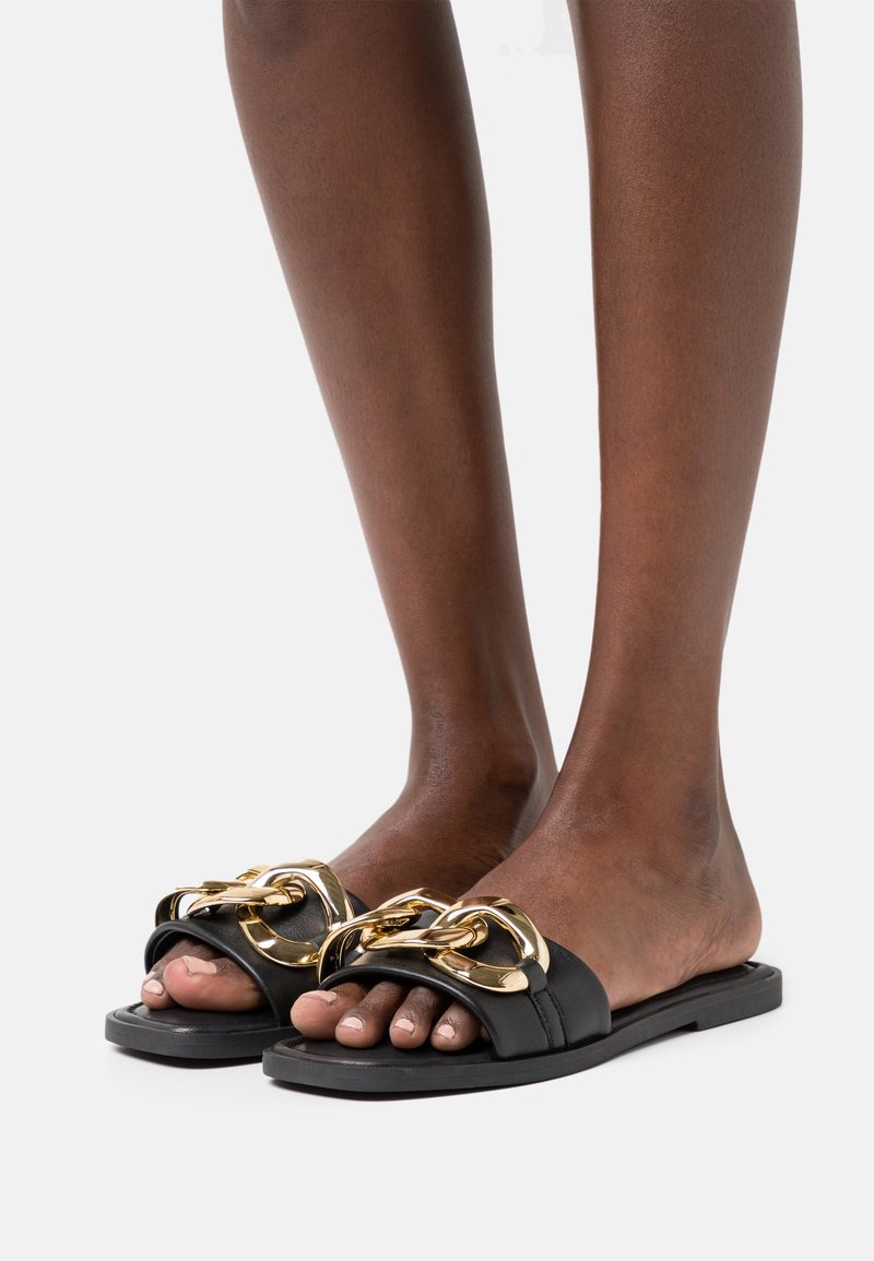Sandales noires à glissière avec une large sangle présentant de grands accents en chaîne dorée. Semelle plate avec une finition lisse et un design à bout ouvert.