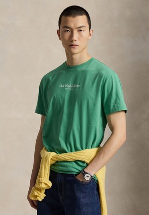 Άνδρας που φοράει πράσινο μπλουζάκι Polo Ralph Lauren, σκούρο τζιν, κίτρινο πουλόβερ δεμένο γύρω από τη μέση και ρολόι χειρός, στέκεται μπροστά σε μονόχρωμο φόντο.