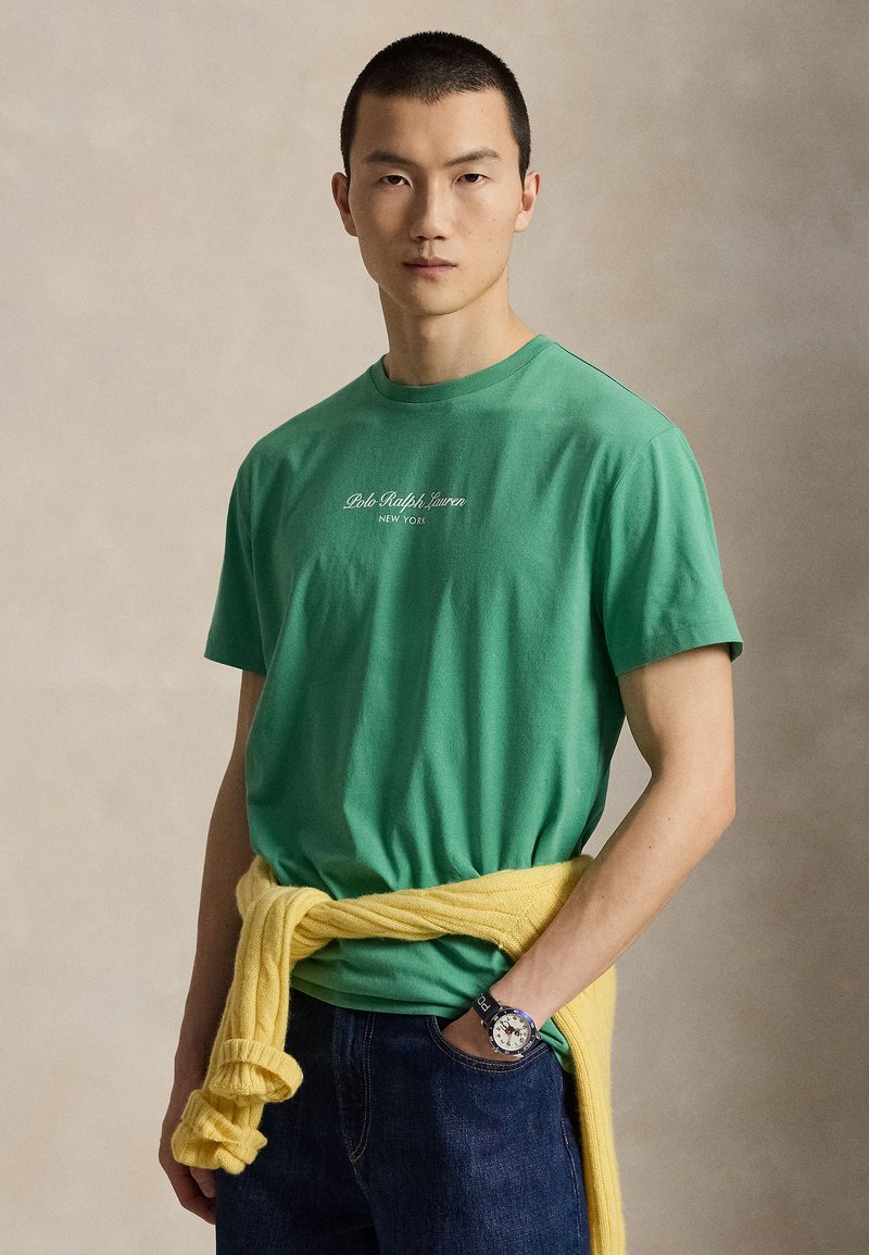 Άνδρας που φοράει πράσινο μπλουζάκι Polo Ralph Lauren, σκούρο τζιν, κίτρινο πουλόβερ δεμένο γύρω από τη μέση και ρολόι χειρός, στέκεται μπροστά σε μονόχρωμο φόντο.