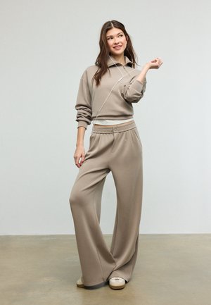 Nuori nainen vaaleanruskeassa rentoutumisvaatetuksessa, leveät housut ja collegepaita, kuulokkeet päässä, seisoo rentoutuneessa asennossa yksivärisen taustan edessä.