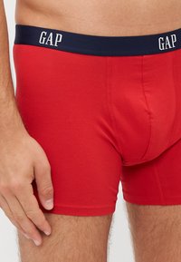 Boxeri scurți roșii din bumbac cu un brâu bleumarin pe care se află logo-ul alb "GAP". Materialul este neted, elastic și ajustat, cu detalii ale cusăturilor.