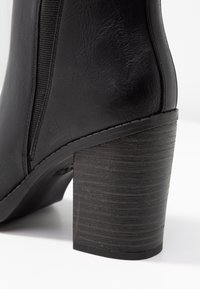 Botín negro con tacón de cuero sintético texturizado. Presenta paneles laterales elásticos y un tacón grueso con textura de madera.