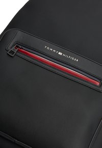 Μαύρο υφασμάτινο σακίδιο πλάτης με κόκκινη, λευκή και μπλε λωρίδα λεπτομέρειας. Διαθέτει φερμουάρ και λογότυπο "Tommy Hilfiger" στο εμπρόσθιο μέρος.