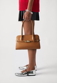 Sac à main en cuir marron avec une forme structurée, des poignées fines et un fermoir en ton doré. Le mannequin porte des chaussures plates argentées et une jupe plissée.