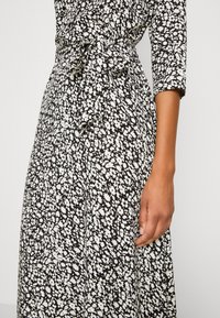 Robe à motifs floraux noir et blanc avec des manches trois-quarts, une ceinture à la taille et une jupe mi-longue. Texture de tissu doux.