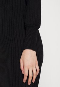 Main droite reposant à côté d'un vêtement à manches longues en maille côtelée noire sur fond blanc.