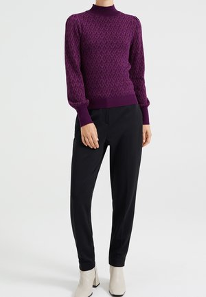 Femme portant un pull à col roulé violet à motifs avec des manches bouffantes, un pantalon noir sur mesure et des bottines blanches, debout droite.