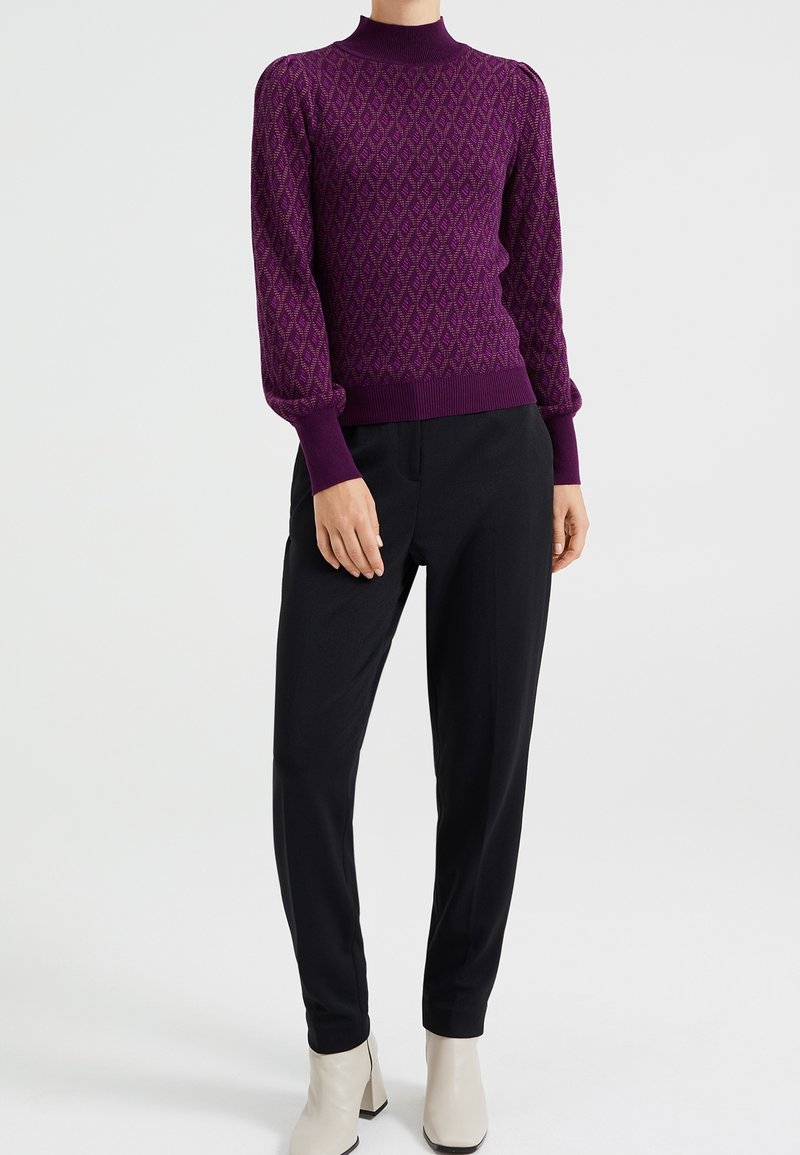 Femme portant un pull à col roulé violet à motifs avec des manches bouffantes, un pantalon noir sur mesure et des bottines blanches, debout droite.