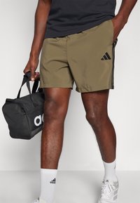 Oliivinvihreät urheilushortsit mustilla sivuilla ja Adidas-logo. Malli pitää mustaa urheilukassia, jossa on valkoiset "adidas"-tekstit.
