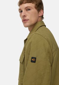 Gequiltede olijfgroene jack met een kraag, borstzak en een zwart label met een oranje logo op de linkerschouder.