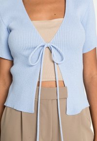 Cardigan de malha canelada azul claro com um decote em V profundo e fecho com laço à frente, combinado com um top cropped bege e calças beges.