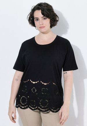 Femme aux cheveux courts, bouclés et foncés, portant un haut noir à manches courtes avec des motifs festonnés découpés et un pantalon beige, debout contre un mur blanc.