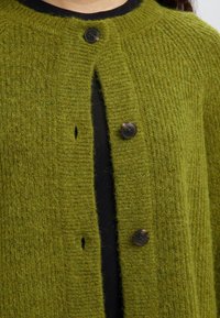 Žalias megztas kardiganas su reljefiniu raštu, apvalia iškirpte ir trimis tamsiomis sagomis priekyje. Matyti juoda marškinėlių apačia.