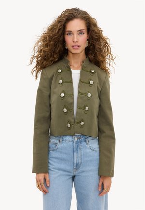 Femme aux cheveux bouclés portant une veste courte vert olive avec des boutons décoratifs et un jean taille haute bleu clair, tournée vers l'avant.