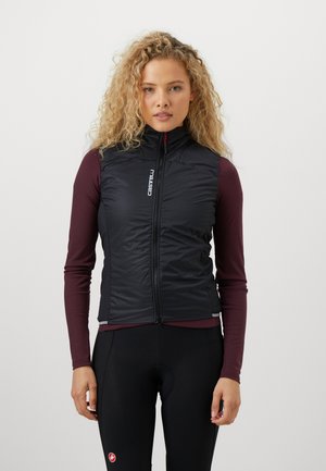 Eine Frau mit lockigem blonden Haar trägt eine schwarze, durchgängige Weste über einem bordeauxroten Langarmshirt und schwarzen Radlerhosen und steht vor einem weißen Hintergrund.