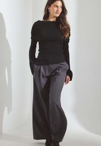 Čierne textúrované tričko s dlhým rukávom a lodičkovým výstrihom, skombinované s vysokými šedými nohavicami so širokými nohami. Model stojí na svetlom pozadí.