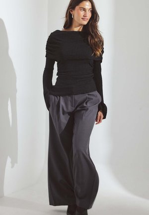 Črna teksturirana dolga majica z jahtno ovratnikom, v kombinaciji s sivo širokokrajnimi hlačami z visokim pasom. Model stoji ob svetli ozadju.